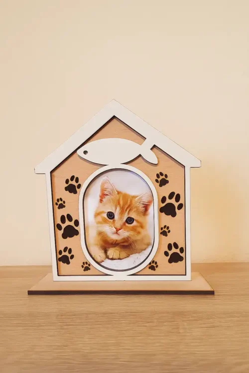 A-cica-1 Natúr fából készült macskaház fényképtartó, amelyben egy aranyos cica képe látható. A keret tetején egy fehér halacska van, oldalán mancsnyomok. A macskaház fényképtartó egy fa asztalon áll.