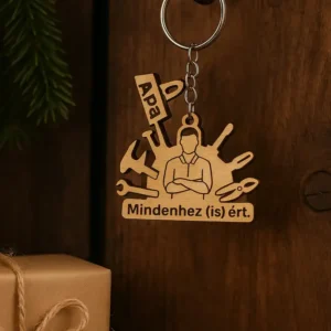 Egyedi gravírozott fa kulcstartó "Apa" és "Mindenhez (is) Ért" felirattal, szerszámokkal. A gravírozott fa kulcstartó egy barna ajándékdoboz mellett fekszik.