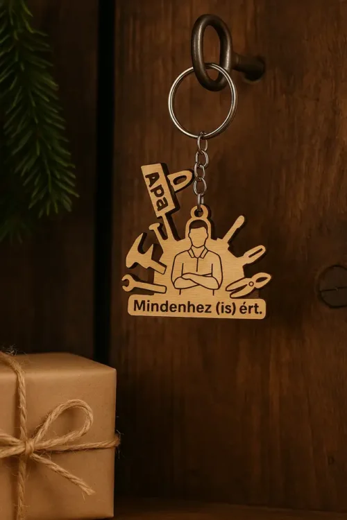 Egyedi gravírozott fa kulcstartó "Apa" és "Mindenhez (is) Ért" felirattal, szerszámokkal. A gravírozott fa kulcstartó egy barna ajándékdoboz mellett fekszik.