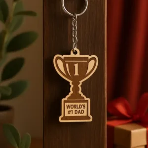 Trófea formájú, lézergravírozott apák napi trófea kulcstartó "World's #1 Dad" felirattal, egy sötét fa felületen lógva. Az apák napi trófea kulcstartó