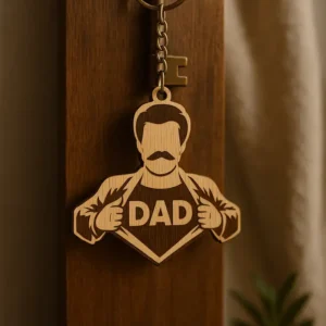 Szuperhős apa alakú, lézergravírozott szuperhős apa kulcstartó "DAD" felirattal, egy sötét fa felületen lógva egy régi kulccsal. A szuperhős apa kulcstartó mellett egy zöld szobanövény látható.