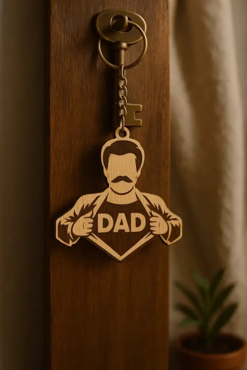 V5 Szuperhős apa alakú, lézergravírozott szuperhős apa kulcstartó "DAD" felirattal, egy sötét fa felületen lógva egy régi kulccsal. A szuperhős apa kulcstartó mellett egy zöld szobanövény látható.