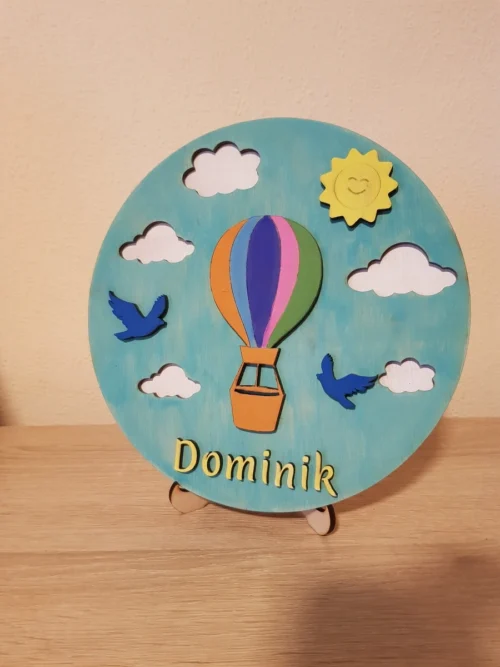 1_result Egyedi hőlégballonos gyerekszoba névtábla "Dominik" névvel, egy fa állványon elhelyezve.