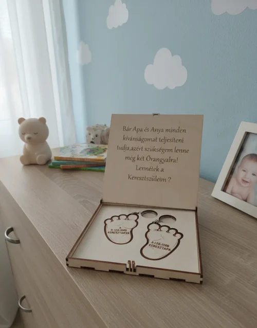 Keresztszülő felkérő ajándékdoboz egy babaszoba komódján, mackó és képkeret mellett.