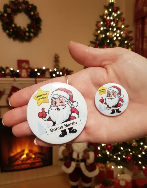 Két különböző méretű, névre szóló mikulás kitűző egy női kézben, karácsonyi háttérrel.