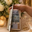fa-eskuvoi-penzatado-tarca-nyitva Kézzel nyitott, fából készült esküvői pénzátadó tárca, amelyben egy húszezer forintos bankjegy látható ajándékként, elegáns esküvői környezetben.